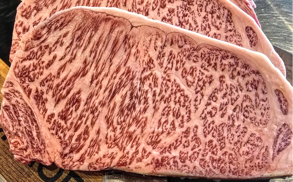 A close-up shot of a thick, beautifully marbled raw New York strip steak. The meat features a dense, intricate pattern of white intramuscular fat scoring perfectly across the vibrant pinkish-red muscle, with a clean strip of white fat along the top edge.