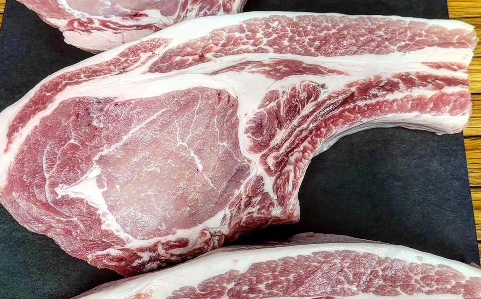 A close-up shot of a thick, raw Duroc pork chop resting on a dark surface. The meat displays a beautiful, distinct marbling of white fat woven throughout the tender pink muscle, featuring a clean layer of white fat along the curved top edge.