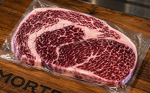 A close-up of a thick, raw, beautifully marbled steak resting on a branded J. Morten's wooden cutting board