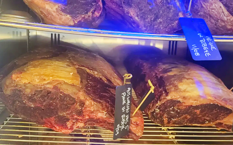 A close-up shot looking inside a dimly lit meat aging room or display case. Several large cuts of USDA Prime beef are resting on wire racks, showcasing a deep, rich purple-red color and a textured layer of dry-aged exterior crust. Small, rustic wooden price tags or markers are stuck into the steaks.