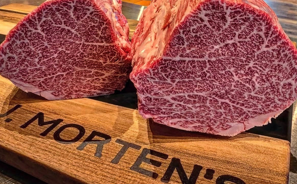 A close-up shot of two thick, heavily marbled cuts of raw Wagyu beef sitting on a dark surface. The meat displays a dense, intricate network of white intramuscular fat (marbling) woven throughout the rich, reddish-pink muscle. In the lower left corner, a portion of a branded wooden box is visible, showing the stamped text "MORTEN'S".
