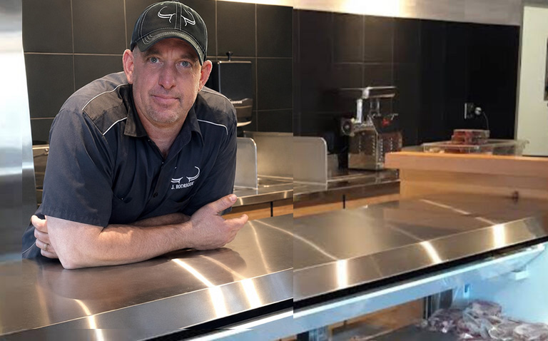 Owner and chef Dan Grilz wearing a baseball cap and branded shirt, smiling behind the clean stainless steel service counter inside J. Morten's Meat Market.