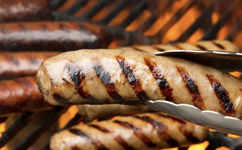 Grilled bratwursts cooking on a grill with metal tongs.