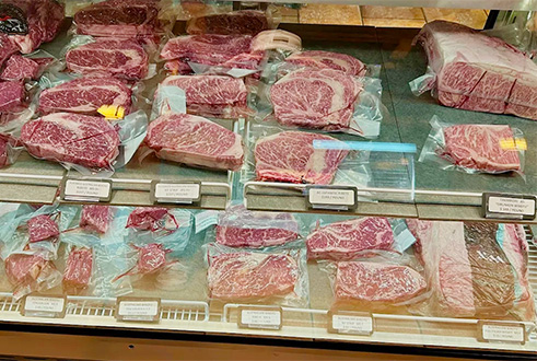A close-up view looking inside a brightly lit butcher's display case filled with various cuts of raw, marbled steaks arranged on small trays.